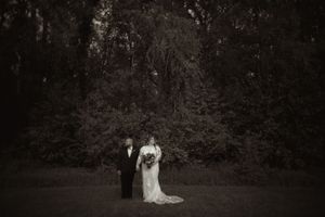 Black and white portrait of bride and groom standing together in front of dark trees at wedding.