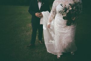 Bride in lace long-sleeve wedding gown holds bouquet beside groom in dark suit on green lawn.