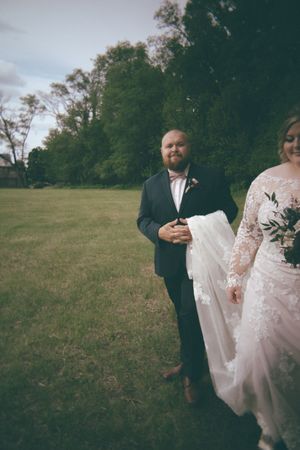 Groom in dark suit carries bride's train as they walk across a grassy field during wedding portraits.