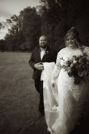 Black and white photo of a groom escorting his bride in a lace gown across an open field.