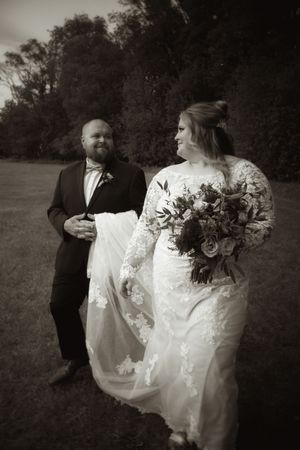 Black and white photo of bride and groom walking outdoors, groom carrying wedding dress train.