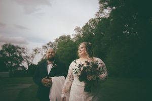 Bride in long lace gown holding bouquet stands beside groom in black suit outdoors.
