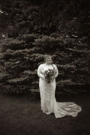 Bride in lace long-sleeve wedding gown holding bouquet stands before evergreen trees in black and white portrait.