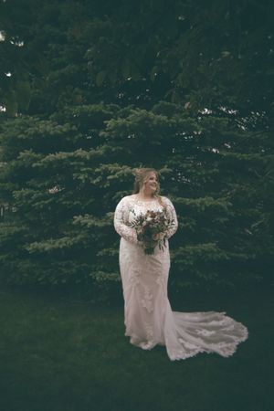 Bride in long-sleeve lace wedding gown holds bouquet standing before lush evergreen trees outdoors.