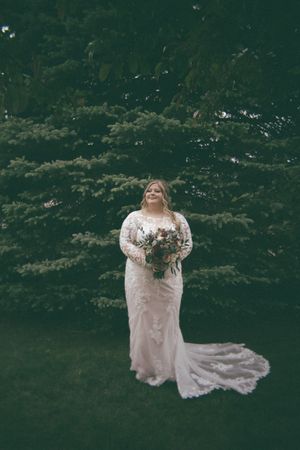 Bride in long-sleeve lace gown with train holds bouquet while standing before tall evergreen trees.