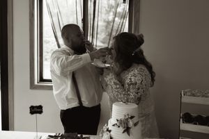 Black and white image of bride and groom sharing cake at wedding reception table near window.