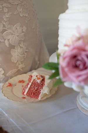 Slice of pink strawberry wedding cake on gold-rimmed plate beside white tiered cake and pink roses.