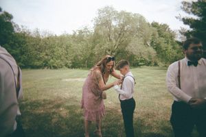 Woman in pink dress helping young boy adjust his suspenders at an outdoor wedding ceremony on a green lawn.