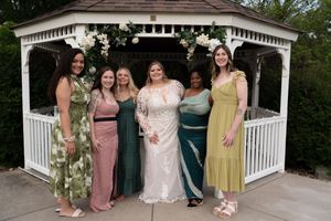 Bride in lace gown poses with five bridesmaids in colorful dresses under white gazebo with flowers.