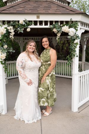 Bride in lace gown poses with guest in green floral dress under white gazebo with white flowers.