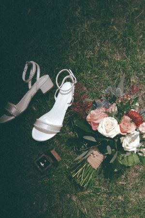 Wedding flat lay featuring block heel sandals, floral bouquet with pink roses, and rings on grass.