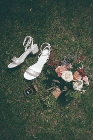 White bridal heels, floral bouquet with pink roses, and a ring box arranged on green grass for a wedding flat lay.