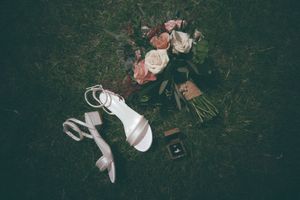 Bridal bouquet of pink and white roses laid on grass beside white strappy heels and a ring box.