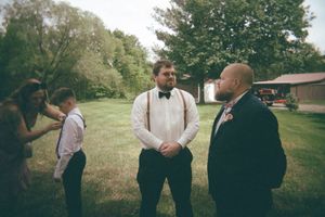 Groom in white shirt and bow tie stands with groomsmen in suspenders on a green lawn outdoors.