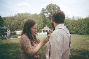 Woman with tattoos adjusting boutonniere on groom's lapel outdoors at wedding venue with gazebo in background.