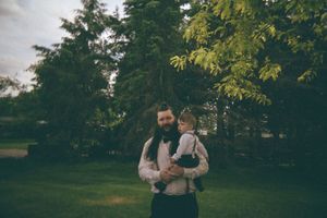 Bearded man in vest and bow tie holds a young child in a lush green park setting.