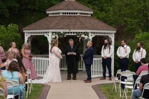 An outdoor wedding ceremony at a white gazebo with bridesmaids, groomsmen, and seated guests.