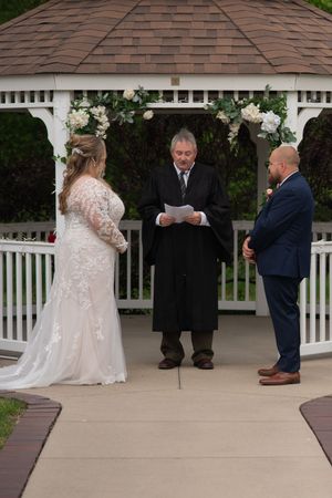 Bride and groom exchange vows before officiant in black robe at flower-adorned white gazebo wedding ceremony.