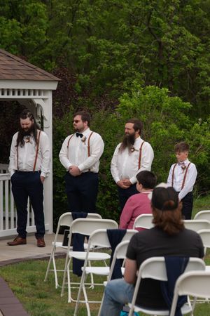 Groomsmen in white shirts and suspenders stand near a gazebo during an outdoor wedding ceremony.