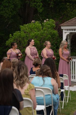 Four bridesmaids in dusty rose dresses holding bouquets stand near a white gazebo outdoors.