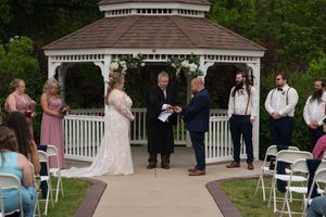 Bride and groom exchange vows at a white gazebo ceremony with wedding party and seated guests.
