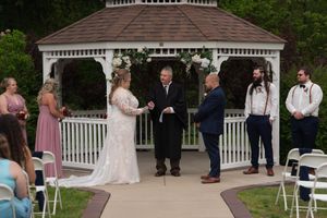 Bride and groom exchange vows at outdoor gazebo ceremony with wedding party and seated guests.