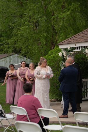 Bride in long-sleeve lace wedding gown reciting vows to groom at outdoor ceremony with bridesmaids in pink dresses.