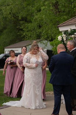 Emotional bride in lace gown reading vows at outdoor wedding ceremony with bridesmaids in mauve dresses behind her.