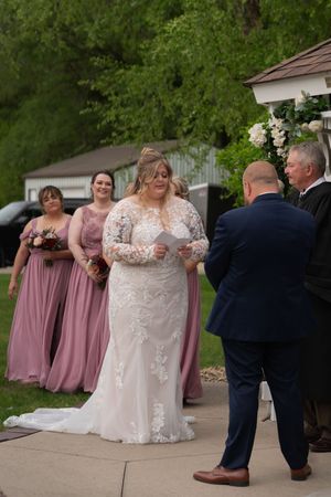 Bride reads vows in lace gown at outdoor wedding ceremony with bridesmaids in dusty rose dresses.
