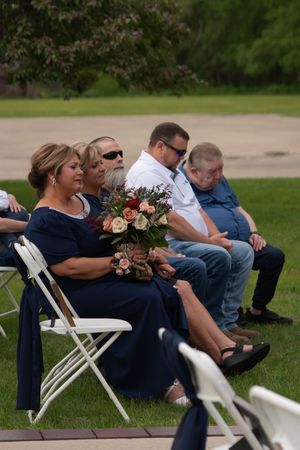 Wedding guests in navy and casual attire sit in white chairs during an outdoor ceremony.