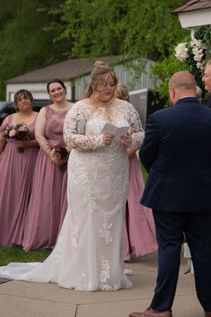 Bride in lace gown reads vows at outdoor ceremony with bridesmaids in dusty rose dresses behind her.