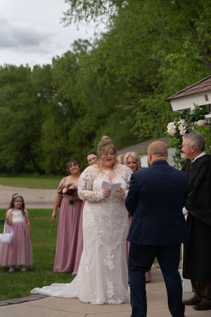 Bride reads wedding vows to groom at outdoor gazebo ceremony with bridesmaids and flower girl.