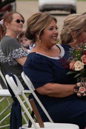 Smiling woman in navy blue dress holds a floral bouquet while seated at an outdoor wedding ceremony.