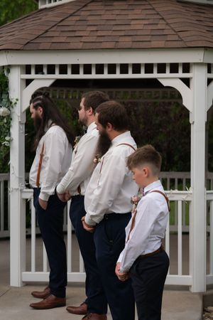 Groomsmen and ring bearer in white shirts with suspenders standing at white gazebo ceremony.