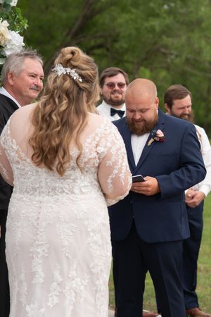 Groom in navy suit reads vows from phone while bride stands facing him during outdoor ceremony.