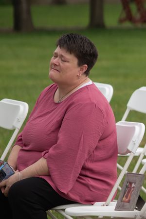 Emotional female wedding guest in pink top sits on white chair during outdoor ceremony.