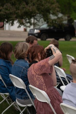 Wedding guest with tattooed arm photographs outdoor ceremony while seated in white folding chair.