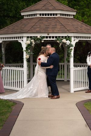 Newlyweds share first kiss at white gazebo altar decorated with white roses and greenery.