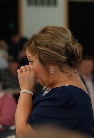 Emotional woman in navy dress wipes tears at wedding reception with elegant updo and earrings.