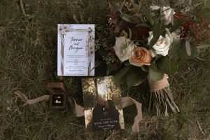 Wedding flat lay with floral invitation, bouquet, rings, and save the date card on grass.