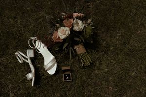Wedding flat lay of white heeled sandals, floral bouquet, and ring box on dark grass.