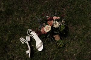 Bridal bouquet with pink and burgundy roses beside white strappy heels on green grass.