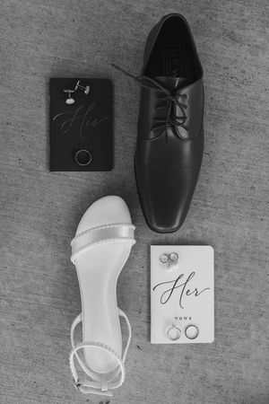 Black and white flat lay of wedding shoes, rings, cufflinks, and His and Her vow books.