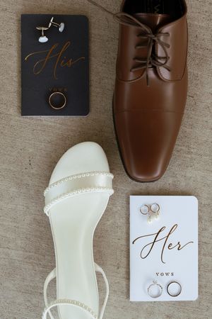 Flat lay of wedding details including his and her vows books, shoes, rings, and earrings.