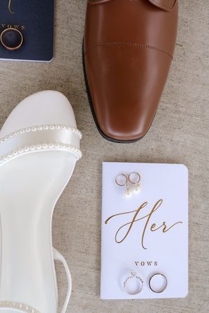 Flat lay of wedding details including white heels, brown dress shoe, vow books, earrings, and gold wedding rings.