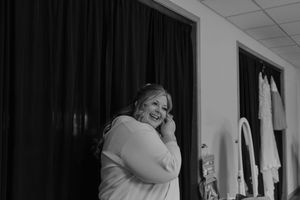 Black and white photo of a smiling bride in a robe touching her face while getting ready for her wedding.