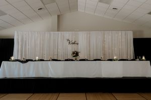 Elegant wedding head table with white draping backdrop, candles, and floral centerpiece in a reception hall.