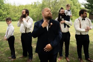 Groom in navy suit adjusts boutonniere surrounded by groomsmen in white shirts and suspenders outdoors.