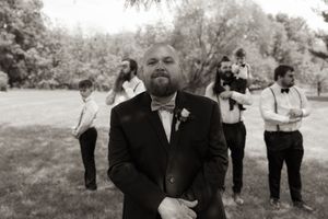 Groom in navy suit with bow tie stands before groomsmen in black and white wedding photo outdoors.