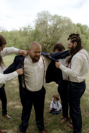 Groomsmen helping bald groom put on navy suit jacket outdoors before wedding ceremony.
